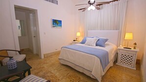 A bedroom with a bed, two bedside tables with lamps, a ceiling fan, a painting on the wall, and a door leading to another room. - Casa del Rocio by Casa de Campo Resort & Villas (La Romana)