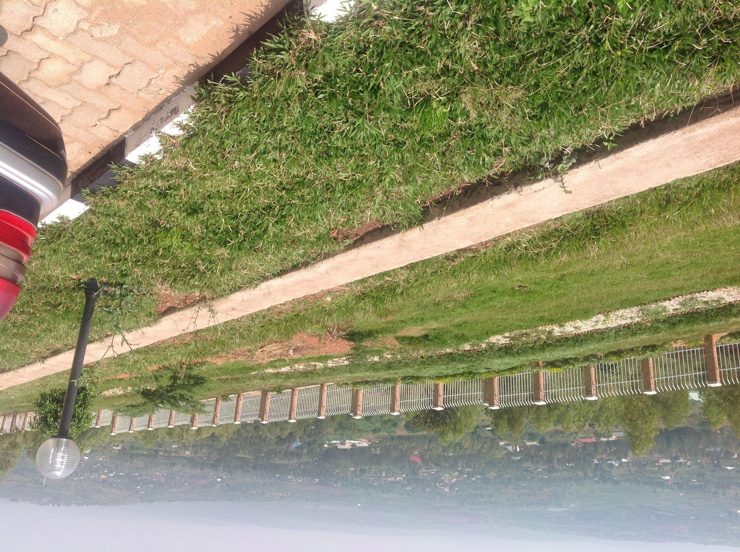 Un área pavimentada con una cerca metálica, una pendiente cubierta de césped y una masa de agua que refleja el entorno.