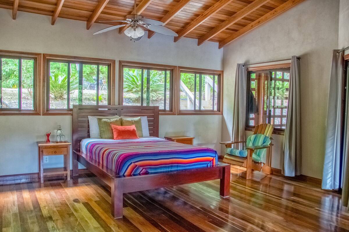 Une chambre à coucher avec un lit en bois, une table de chevet, une chaise et de grandes fenêtres donnant sur un paysage verdoyant.