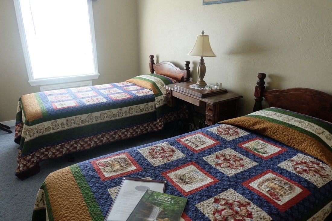 A room with two beds, each covered with a quilt, a nightstand with a lamp, and a window providing natural light.