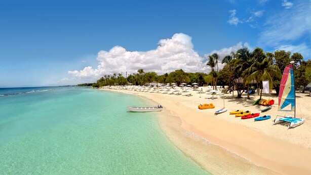 A beach with clear turquoise water, a row of colorful kayaks, and a sailboat.