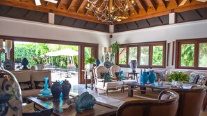 A spacious living area with a high ceiling, wooden beams, and large windows offering a view of greenery outside.