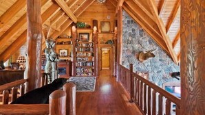 A wooden interior with a high ceiling, a stone wall with a mounted deer head, and a wooden staircase.