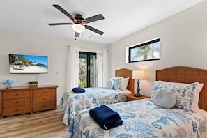 A hotel room with a bed, a nightstand, a TV, a ceiling fan, and a window with a view of palm trees.
