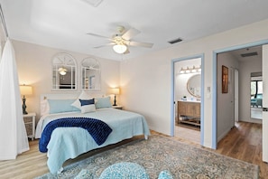 A bedroom with a bed, two bedside tables, a ceiling fan, a large window with a view, and a bathroom with a mirror and sink.