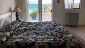 A bedroom with a bed, a nightstand, a lamp, a window, and a view of the ocean.
