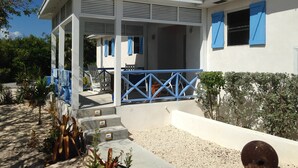 A white building with blue shutters, a covered porch with white chairs, and a gravel pathway leading to the entrance.