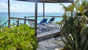 A wooden deck with two blue lounge chairs under a pergola, overlooking a beach with clear turquoise water.