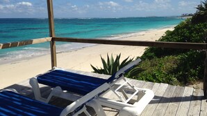 A wooden deck with a blue lounge chair and a white chair overlooking a beach with clear turquoise waters and a sandy shore.