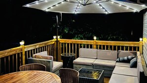 An outdoor deck with wooden flooring, a dining table, wicker chairs, a sofa, and an umbrella with lights. - Tranquilla Villa. Bi Level Deck, HOT TUB, fire pit, grill, Movie & Game Rooms! (Tobyhanna)
