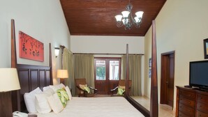 Quarto de hotel com uma cama grande, duas poltronas, uma televisão, um espelho e teto de madeira.