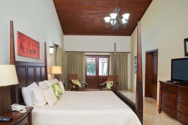 A hotel room with a large bed, two armchairs, a TV, a mirror, and a wooden ceiling.