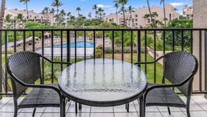 Ein Balkon mit einem Glastisch und zwei Stühlen, der auf einen Pool und einen Resortbereich mit Palmen und Gebäuden blickt.