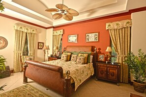 Une chambre spacieuse avec un grand lit, une table de chevet en bois, une commode et un ventilateur de plafond.
