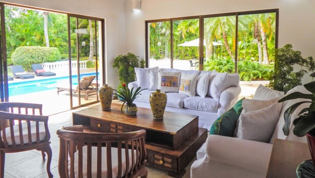 A spacious living area with a white sofa, wooden coffee table, and two chairs. Large windows offer a view of a swimming pool and lush greenery. - Villa Toscano by Casa de Campo Resort & Villas (La Romana)