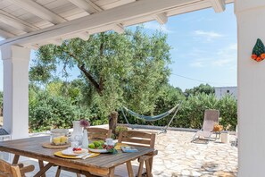 Eine Terrasse im Freien mit einem Holztisch, gedeckt für ein Frühstück, einer Hängematte und Blick auf Olivenbäume und Grünpflanzen.