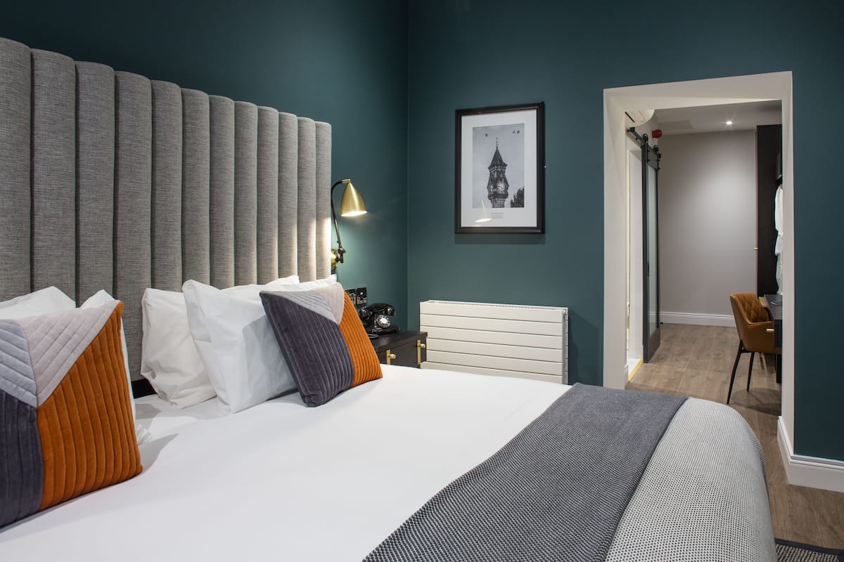 Una habitación de hotel moderna con una cama grande, una mesita de noche con lámpara, un cuadro enmarcado en la pared y una puerta que da a un baño.