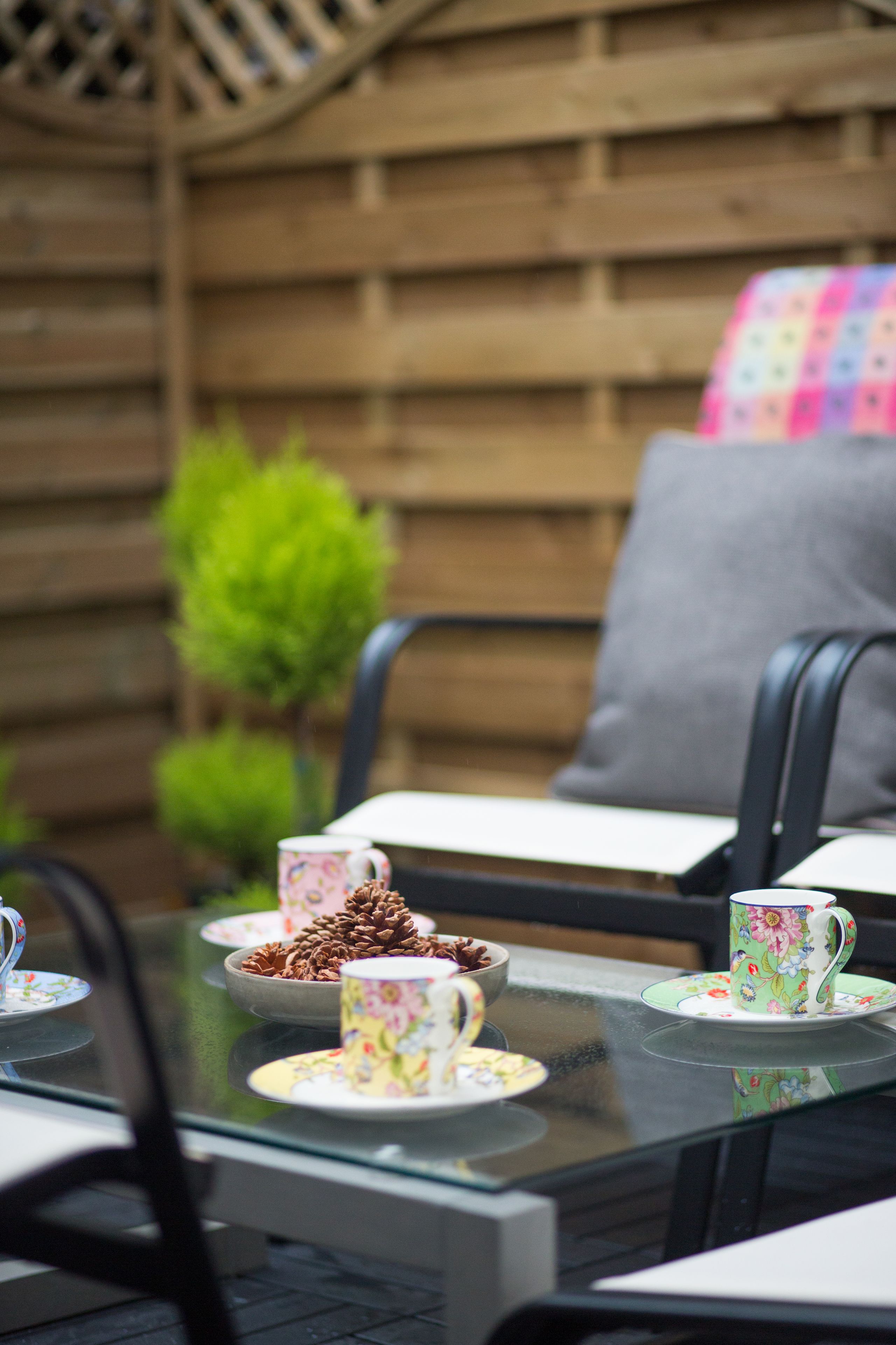 A glass-top table with floral-patterned cups and saucers, a bowl of pinecones, and a small potted plant in a wooden garden setting.