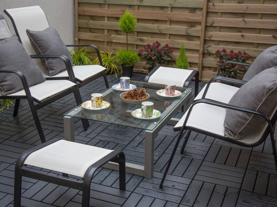A patio with a glass-top table, surrounded by four chairs with cushions. There are potted plants and a wooden fence in the background.
