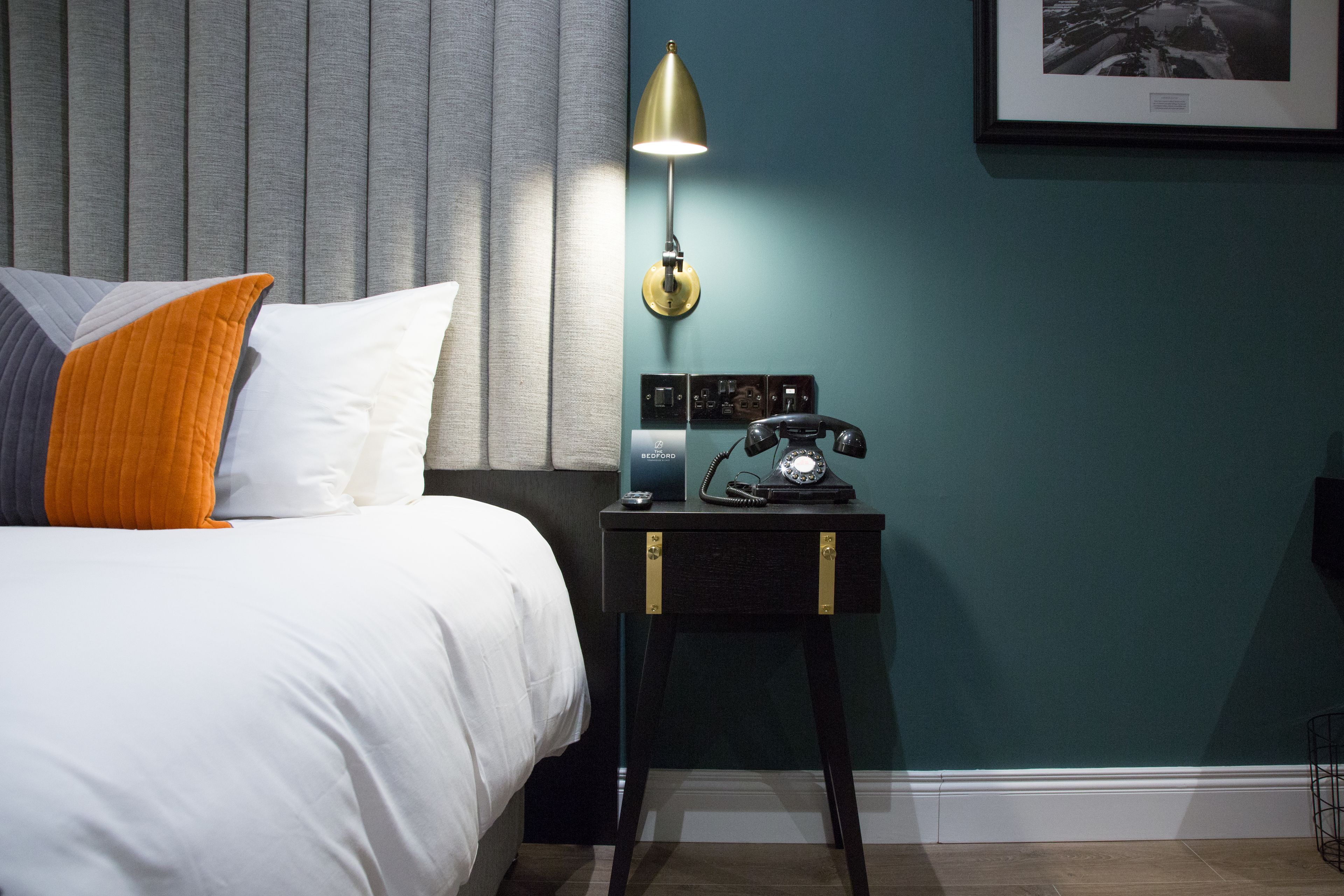 A neatly made bed with white and orange pillows, a vintage-style telephone on a small table beside it, and a wall-mounted picture frame.