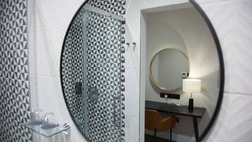 A bathroom with a geometric patterned wall, a glass shower enclosure, a round mirror, a bedside table with a lamp, and a chair.