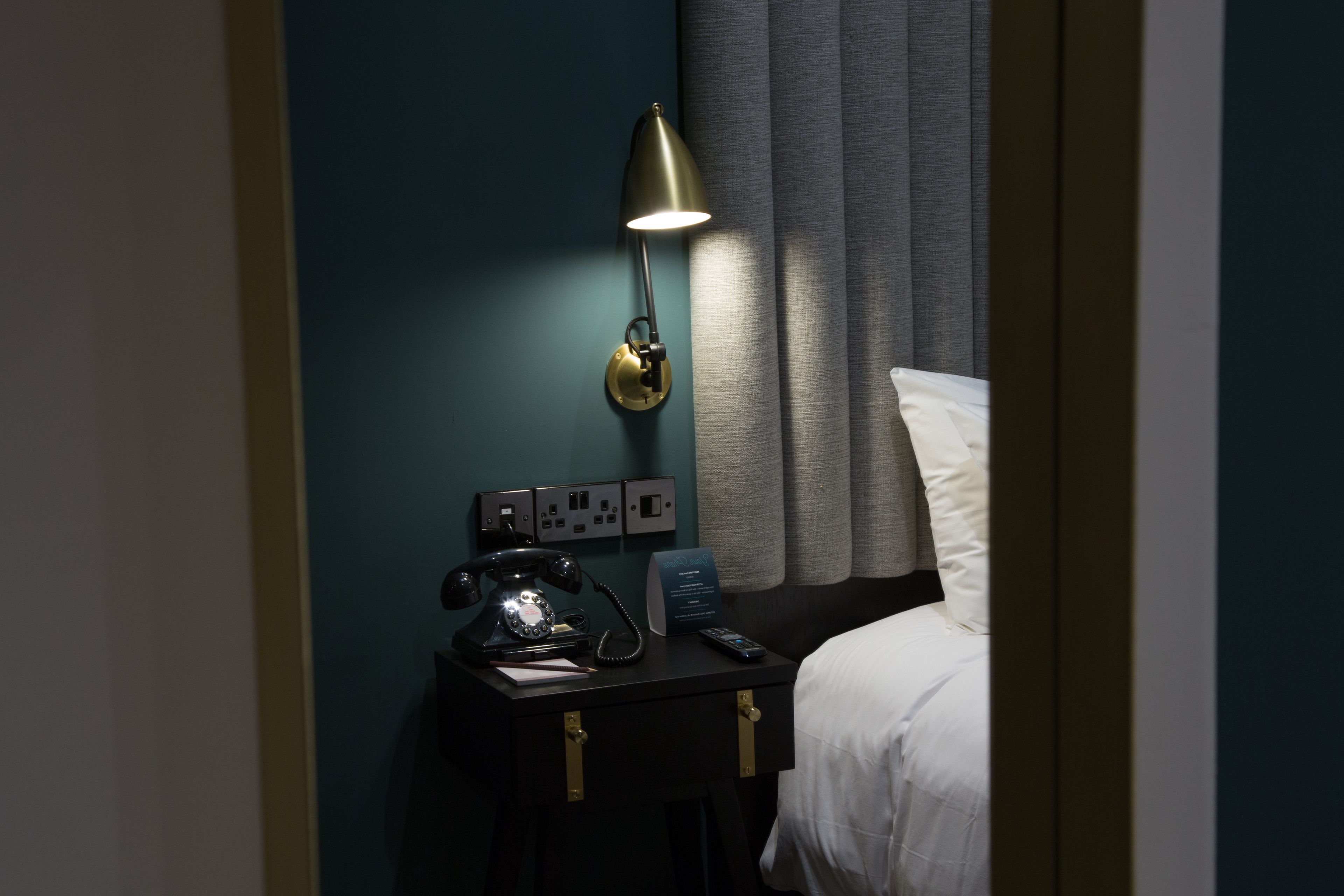 A hotel room with a bedside lamp, a telephone, and a control panel on the wall.