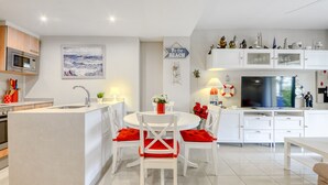 Una cocina moderna con armarios blancos, una isla central y una zona de comedor con sillas blancas y cojines rojos.