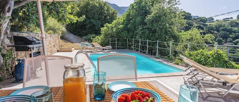 A poolside table with a bowl of strawberries, a pitcher of orange juice, and a glass of water, overlooking a swimming pool and lush greenery.