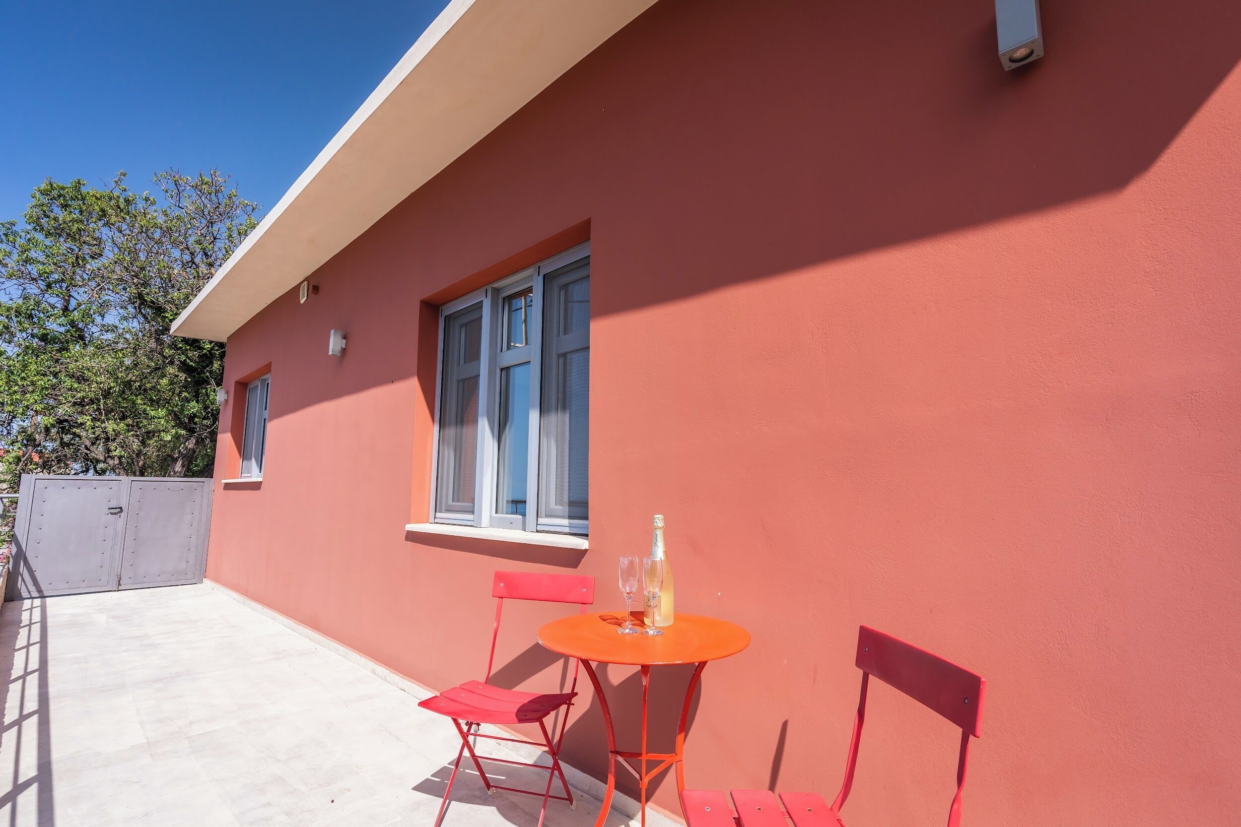 A red building with a window, a table with a bottle and glasses, and two red chairs.