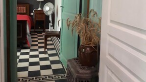 Una habitación con piso a cuadros, una cama con cubre cama rojo, una lámpara, un ventilador y un florero con plantas secas.