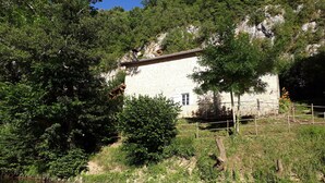 Un edificio de piedra con una ventana pequeña, rodeado de abundante vegetación y un río tranquilo.