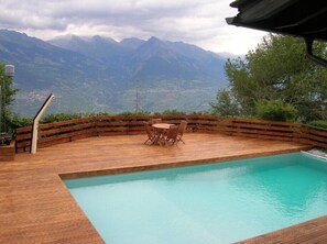 Une terrasse en bois avec une piscine et une table entourée de chaises, offrant une vue sur un paysage montagneux.