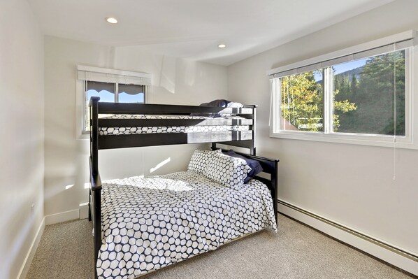 A bunk bed with a patterned bedspread, a window with a view of trees, and a ceiling light.