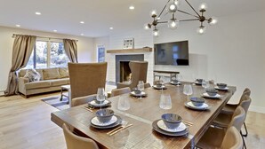 Un comedor con una larga mesa de madera dispuesta para una comida, rodeada de sillas, una chimenea y un televisor en la pared.