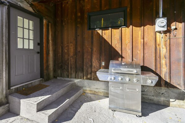 A wooden cabin with a metal grill outside, steps leading to a door, and a power outlet on the wall.