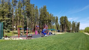 Ein Spielplatz mit verschiedenen Spielgeräten, umgeben von einer Rasenfläche und hohen Bäumen.