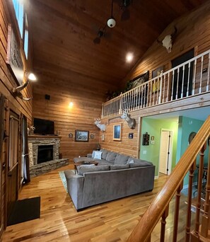 A cabin interior with a stone fireplace, a gray sectional sofa, wooden floors, a balcony with deer antlers, and a staircase.