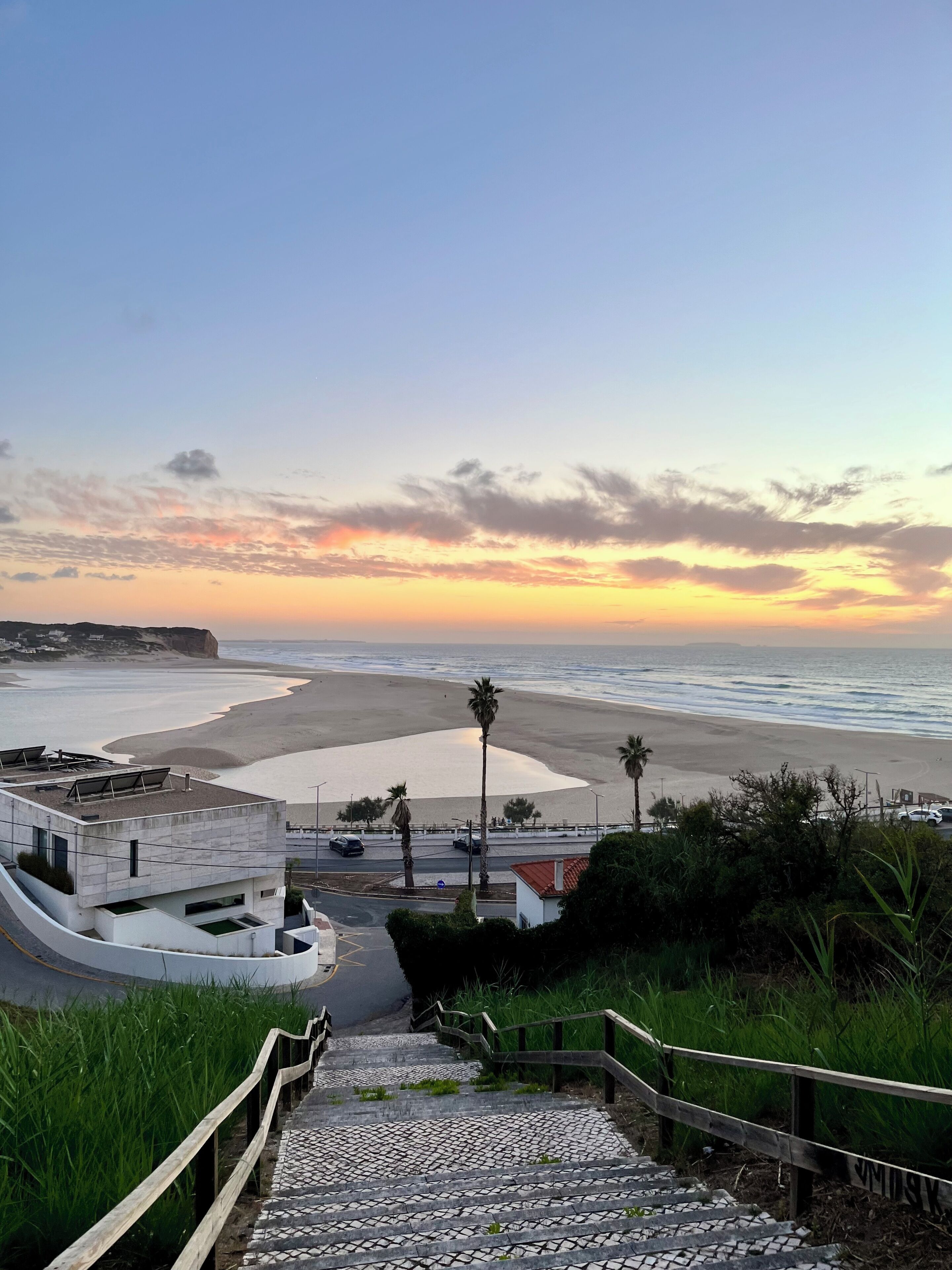 A coastal view with a paved pathway leading to a beach, palm trees, and a modern building with solar panels.