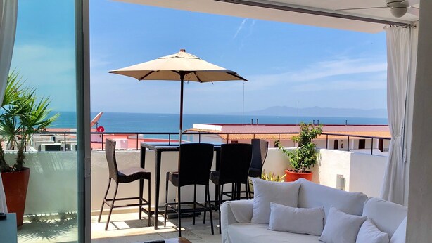 A balcony with a white sofa, a table, and chairs, overlooking a scenic view of the sea and buildings. - COOL 1 bedroom large condo with spa like bath.... enjoy!! (Col. Em. Zapata, Puerto Vallarta)