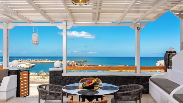 Un espacio al aire libre techado con vista al mar, una mesa redonda con fruta y bebidas, y sillas de mimbre.