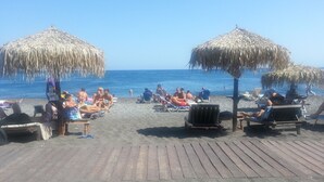 Una playa con sillas de madera y sombrillas de paja, un cielo azul despejado y el océano al fondo.
