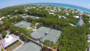 An aerial view of a residential area with multiple tennis courts surrounded by dense greenery and a clear view of the ocean.