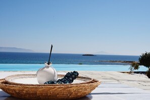 Uma cesta de vime com um vaso branco e uvas em um terraço com piscina e vista para o oceano.