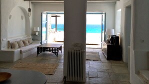 A room with a stone floor, a sofa, a table, and a view of the sea through an open door. - Villa-waterfront, infinity pool-splendid views-luxurious amenities  (PAROS)