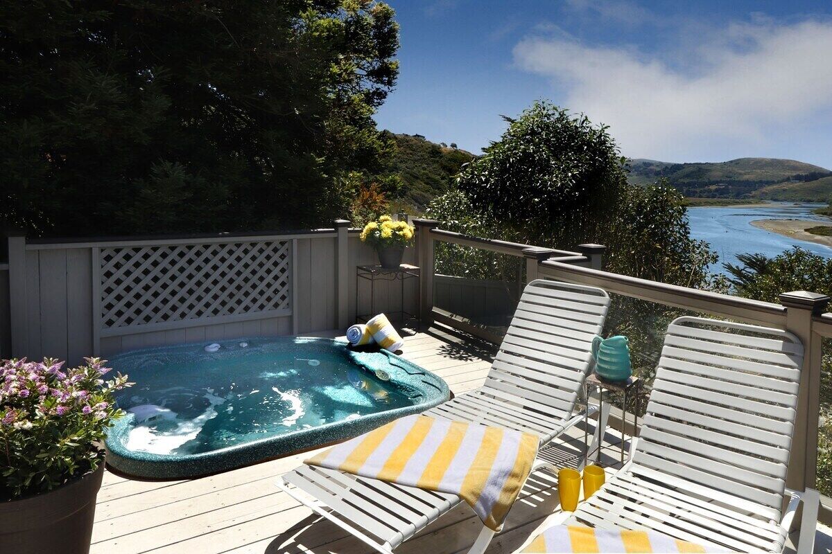 A hot tub on a wooden deck with white lounge chairs and a view of a river and hills.