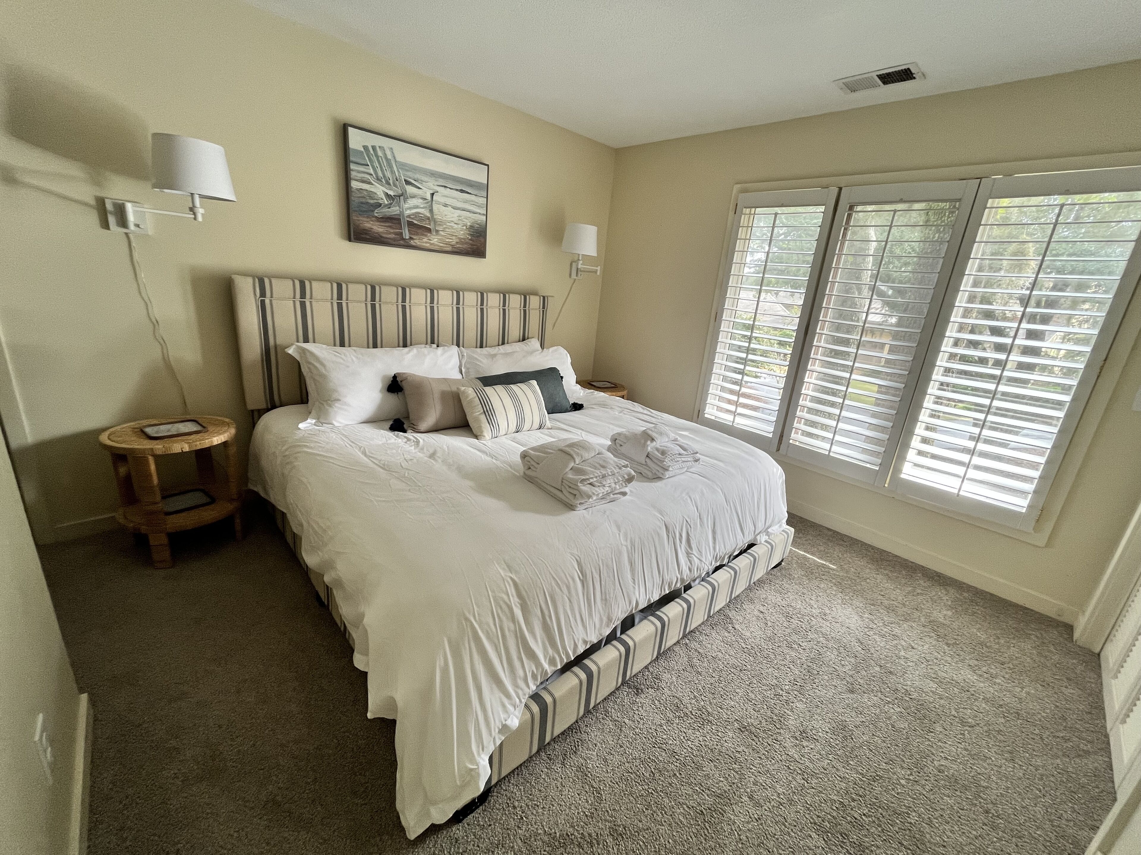 A bedroom with a large bed, a bedside table, a lamp, a painting, and a window with blinds.