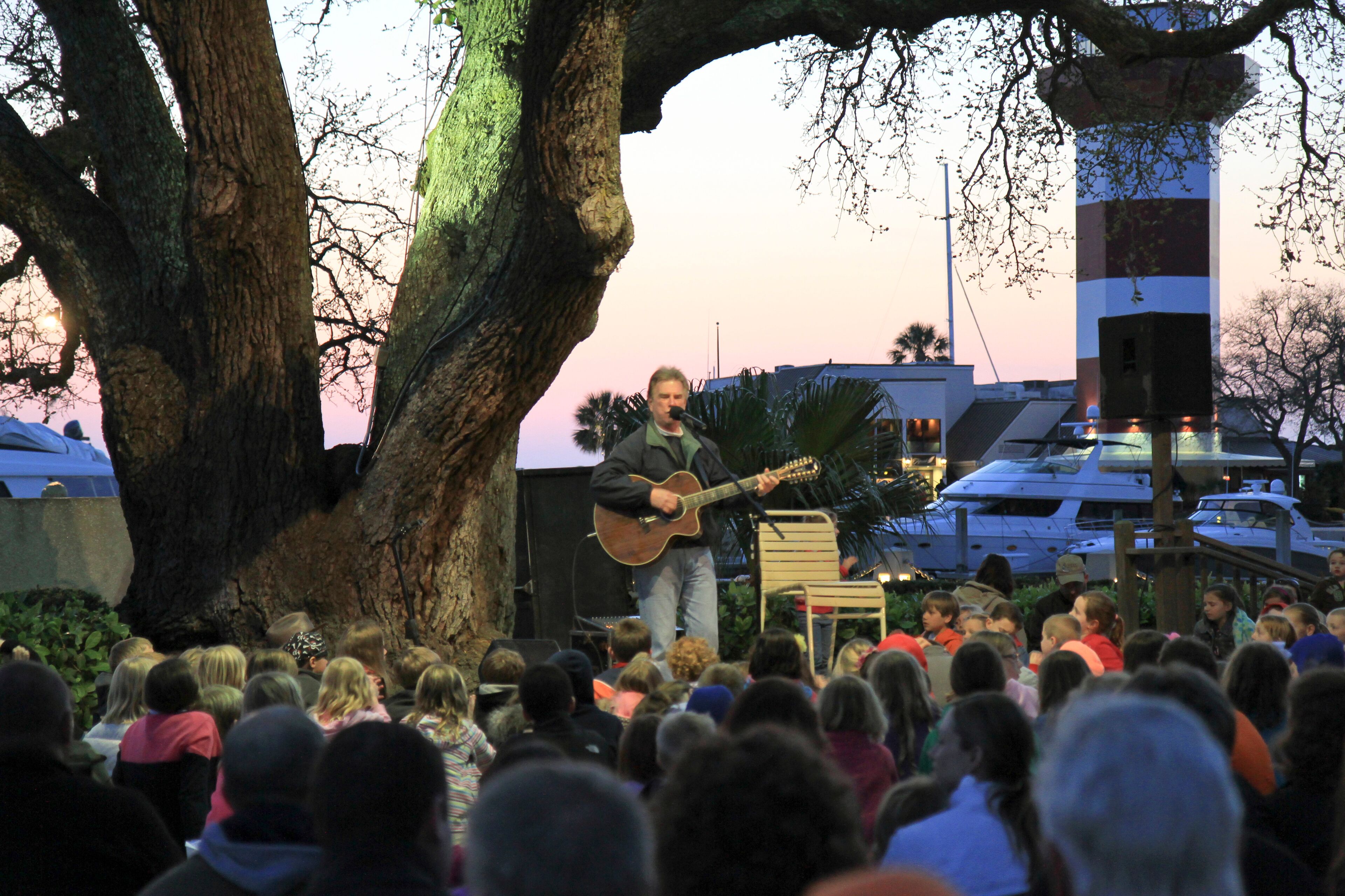 A person playing guitar on a stage with an audience in front of a tree and buildings in the background.