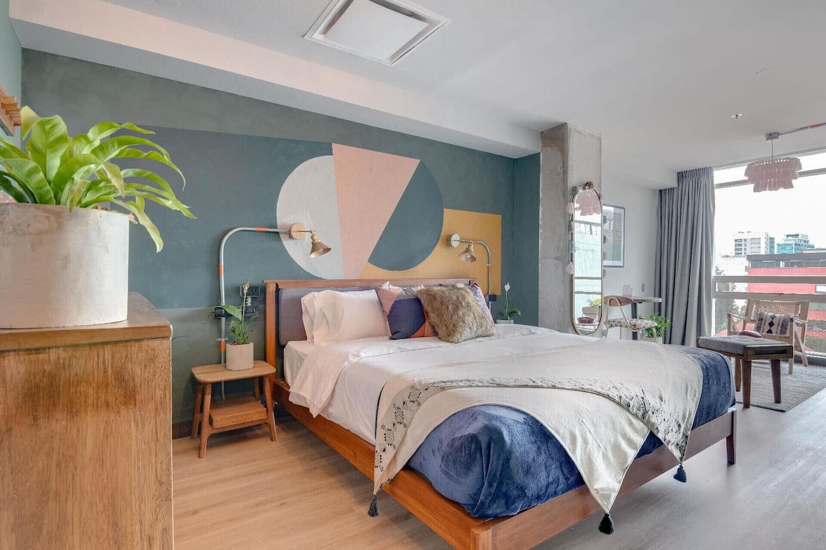 Et moderne soveværelse med en stor seng, en potteplante, et skrivebord og udsigt over byen.
