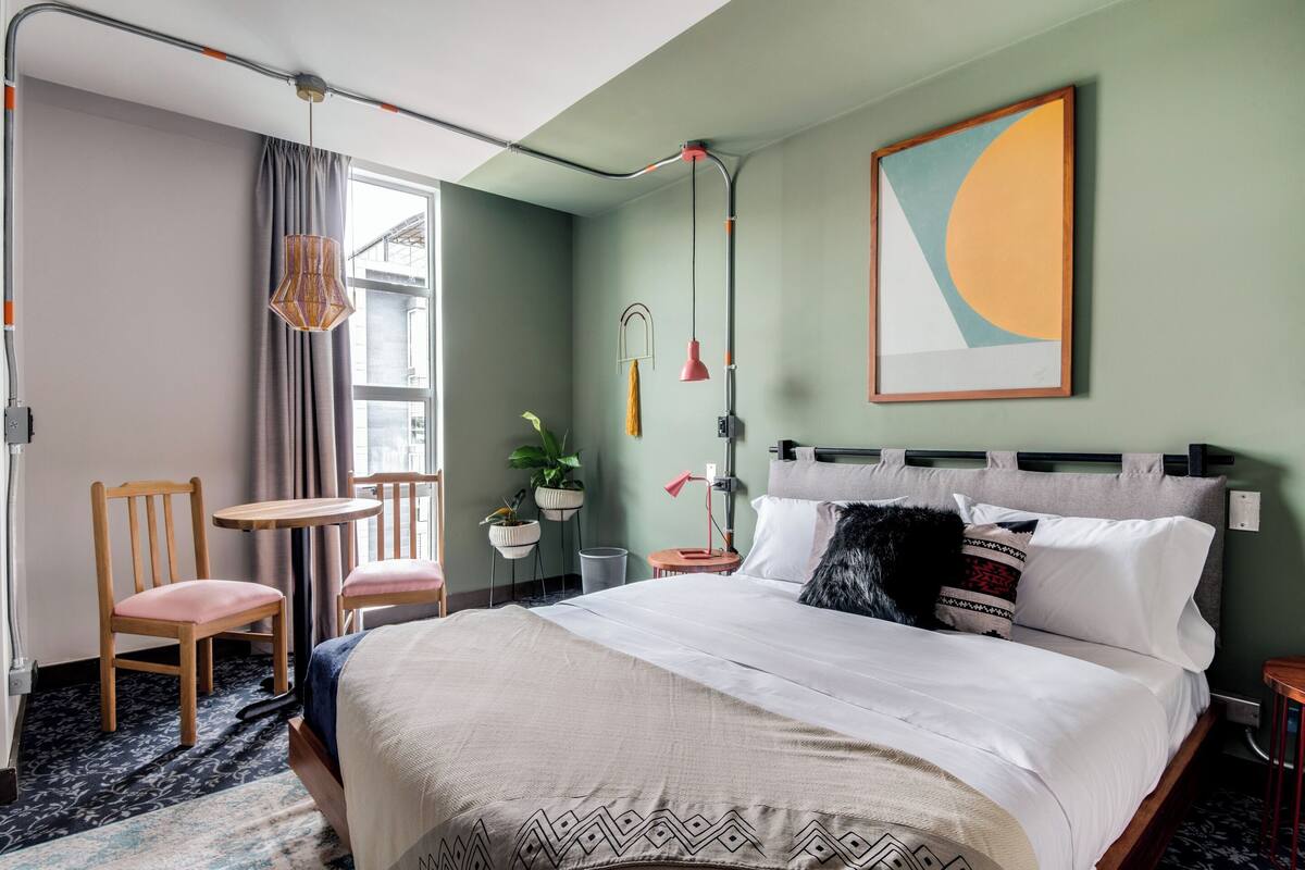 Et hotelværelse med seng, to stole, et lille bord, en potteplante og et maleri på væggen.