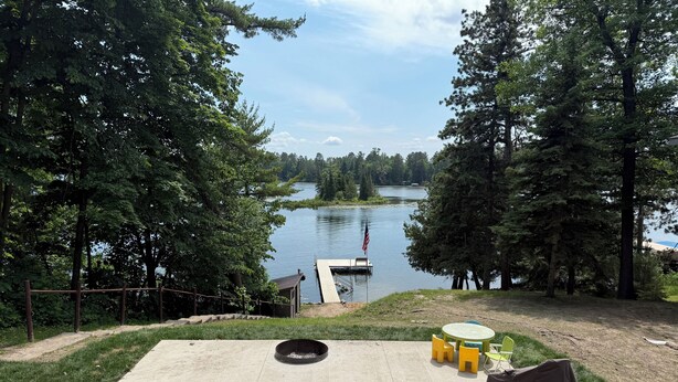 Un patio junto al lago con dos bancos de metal, un pequeño hogar para fogata y un muelle que se adentra en el agua.